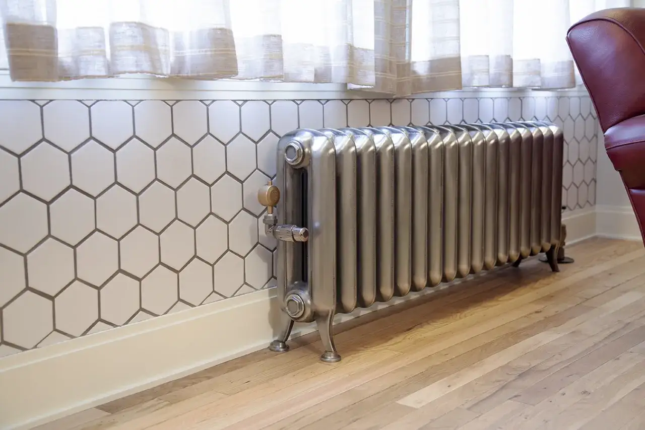 Stary kaloryfer żeliwny, który waży sporo, stoi na drewnianej podłodze przy ścianie z heksagonalnymi płytkami.