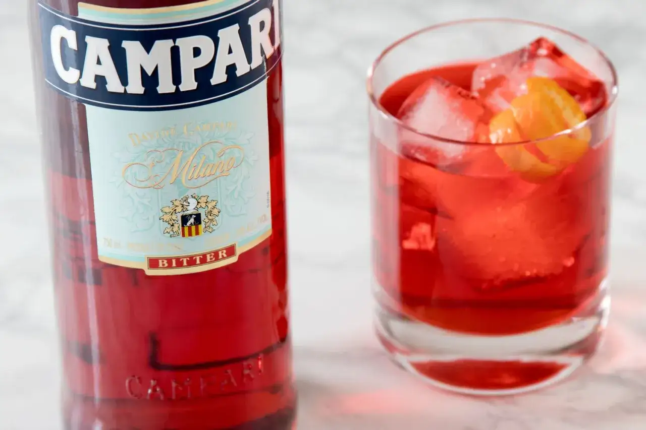 Butelka Campari i szklanka z czerwonym drinkiem, lodem i skórką pomarańczy. Idealny aperitif, który może być bazą do drinków z Aperolem.