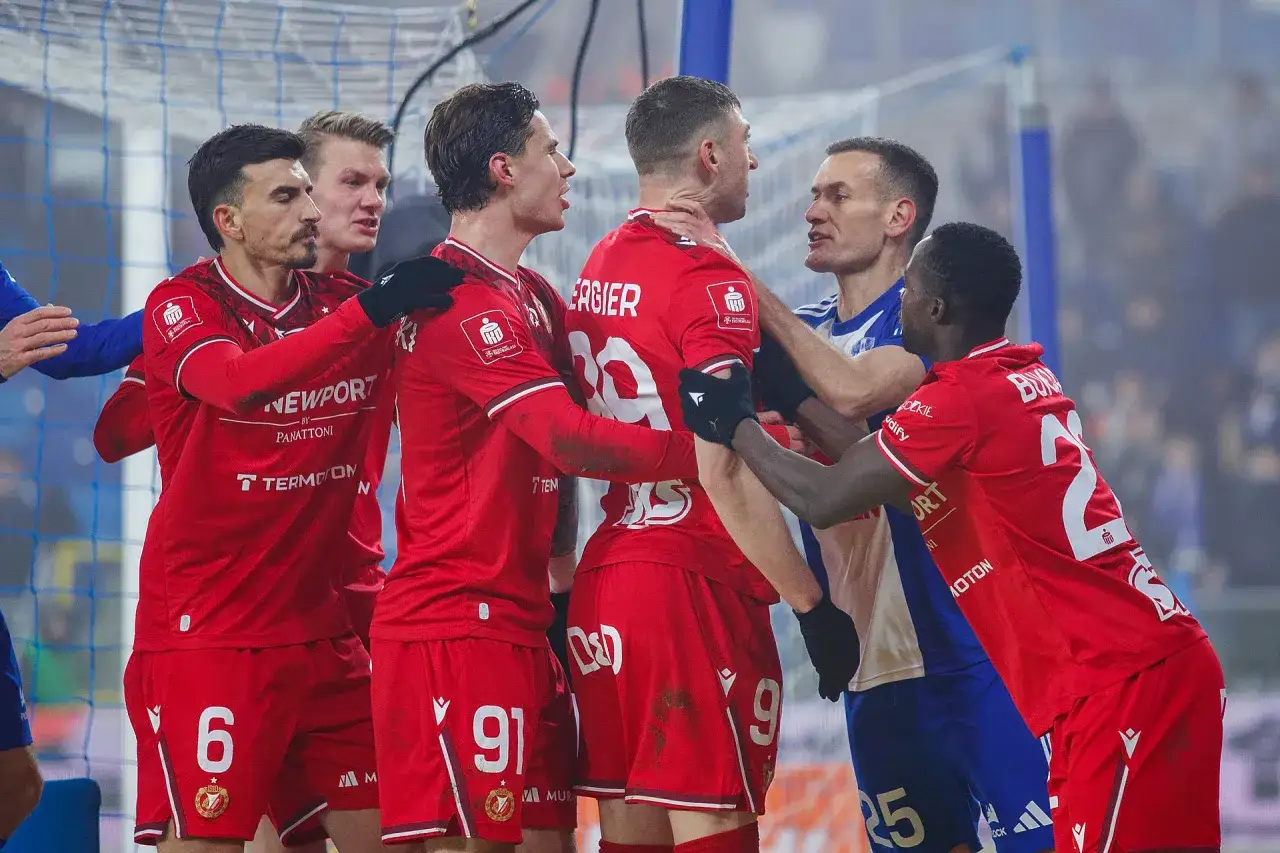 Piłkarze w czerwonych strojach kłócą się z zawodnikiem w niebieskiej koszulce.