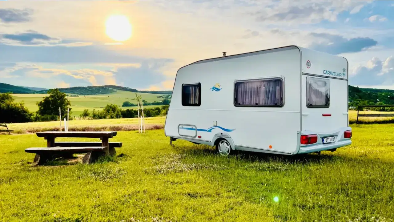 Przyczepa Caravelair zaparkowana na łące o zachodzie słońca. W tle malownicze wzgórza i pola.