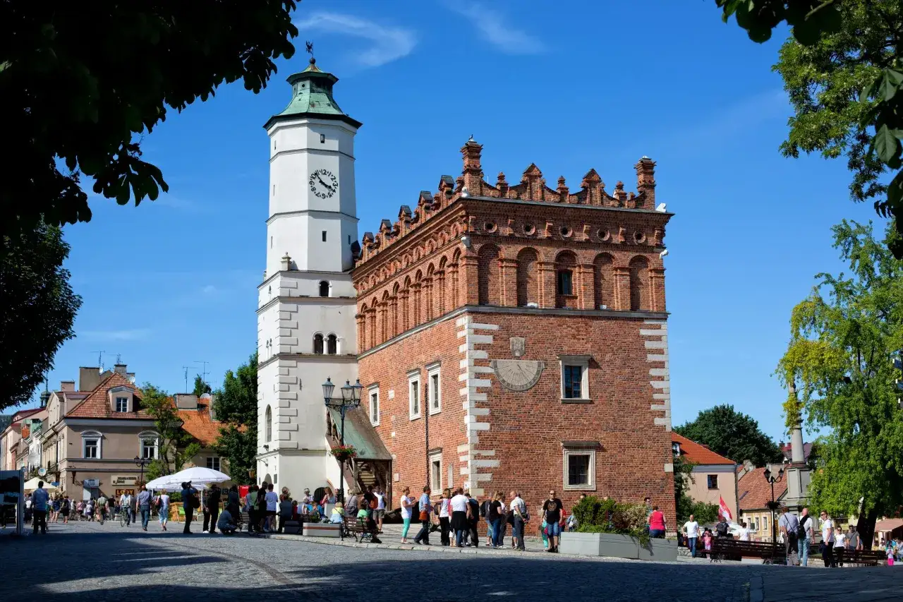 Ratusz w Sandomierzu z zegarem i zegarem słonecznym, na tle błękitnego nieba i drzew.