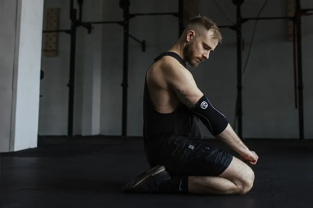 Mężczyzna w czarnym podkoszulku i spodenkach klęczy na siłowni, opierając się na łokciach.