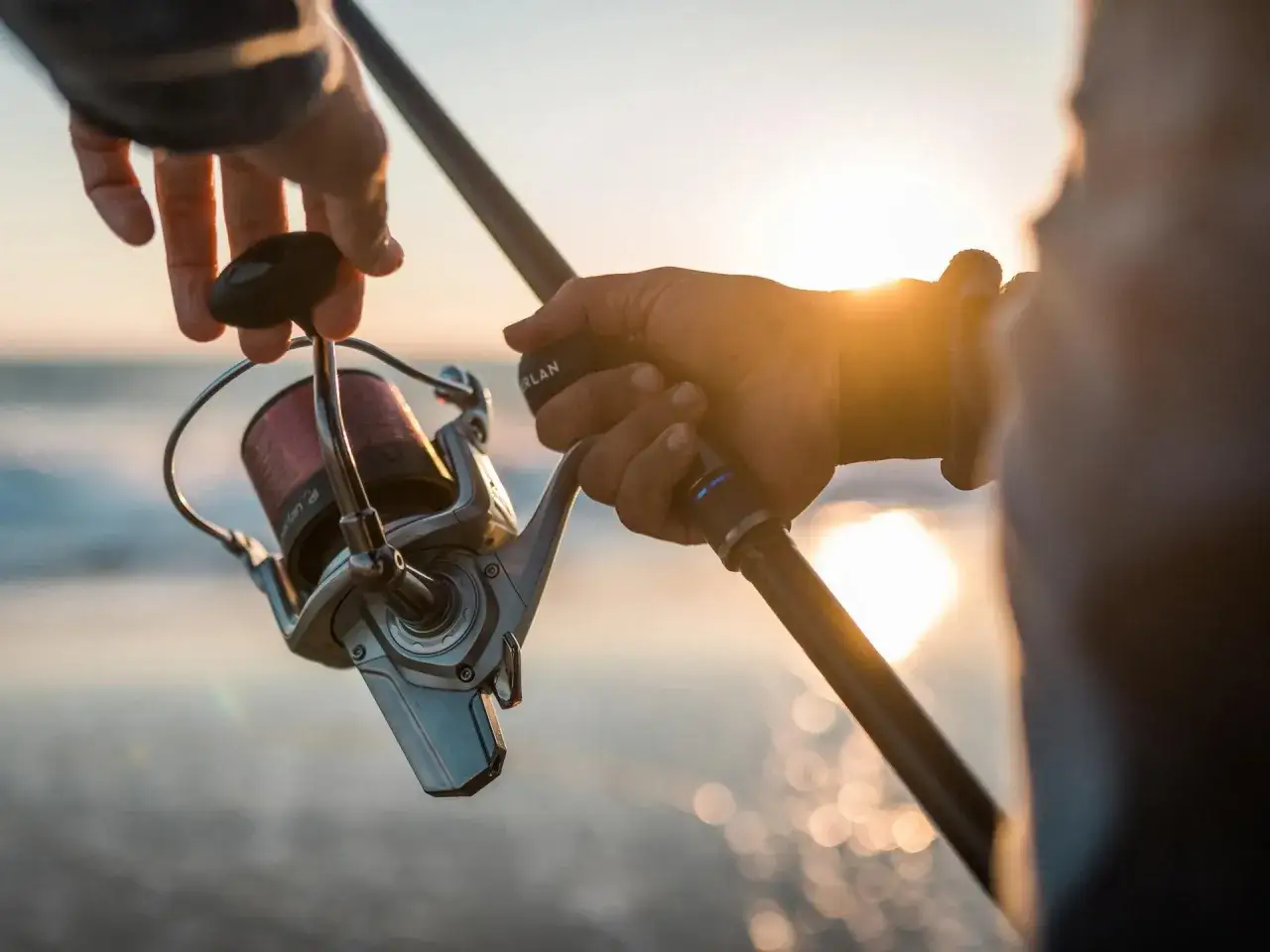 Wędkarz trzyma mocny kołowrotek i wędkę, gotowy na połów suma z gruntu. Słońce zachodzi nad wodą.