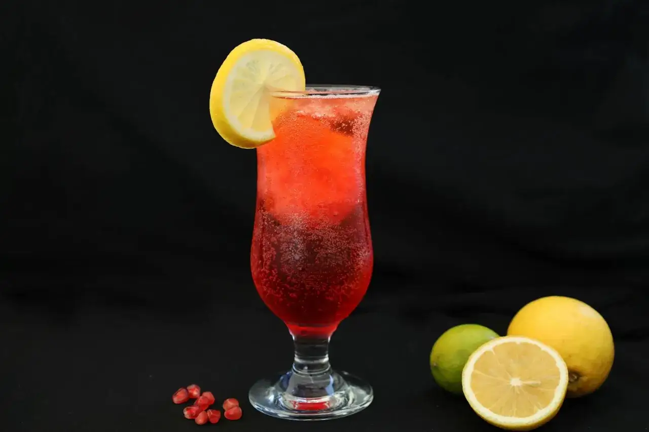 Orzeźwiający drink z granatem, ozdobiony plasterkiem cytryny. Obok leżą świeże owoce: cytryna i limonka.