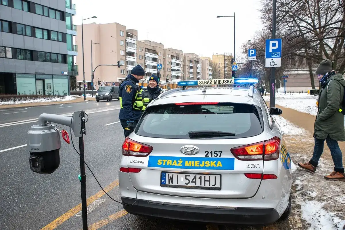 Strażnicy miejscy w Warszawie przy radiowozie i kamerze monitoringu. Dbają o czystą strefę i porządek.
