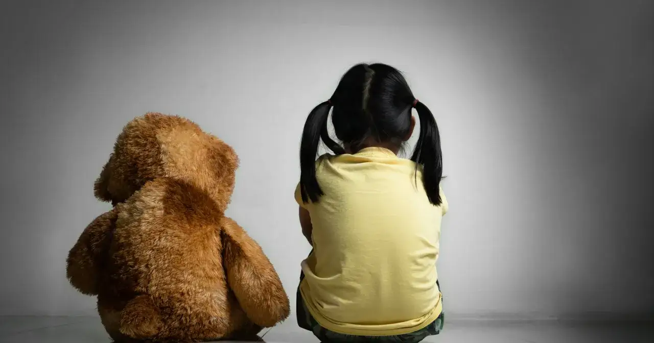 Dziewczynka z kitkami siedzi tyłem do widza obok pluszowego misia.