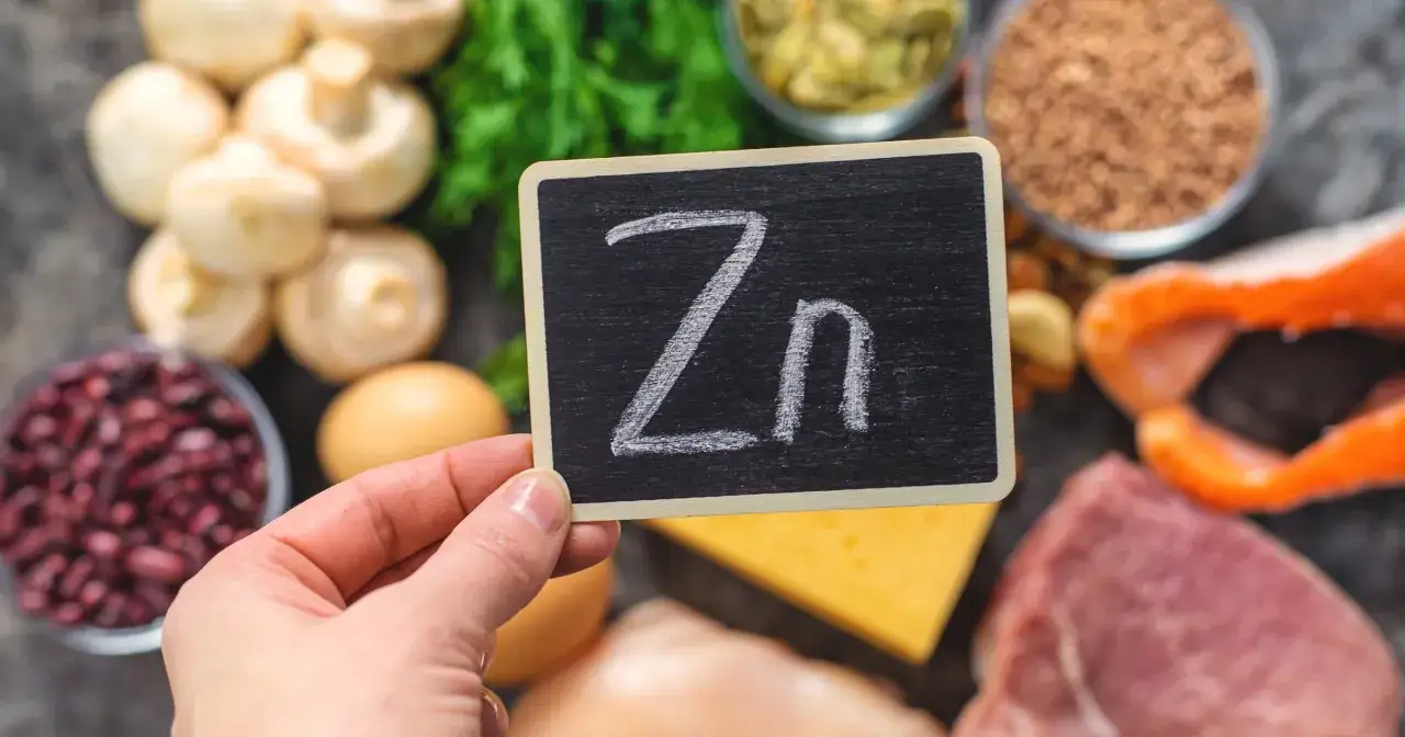Tabliczka z napisem "Zn" na tle produktów bogatych w cynk: mięso, ryby, fasola, grzyby, jajka, ser, nasiona.