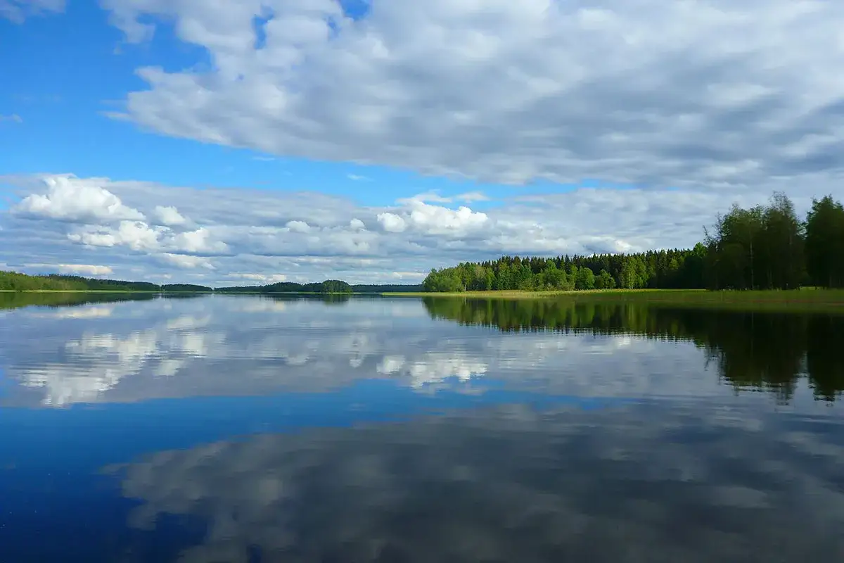 Spokojne jezioro na pojezierzu fińskim, odbijające chmury i zielone lasy.