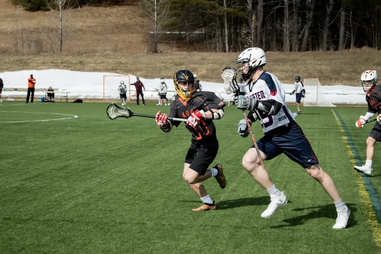 Dwóch zawodników w akcji podczas meczu lacrosse. Jeden biegnie z pałką, drugi go ściga.