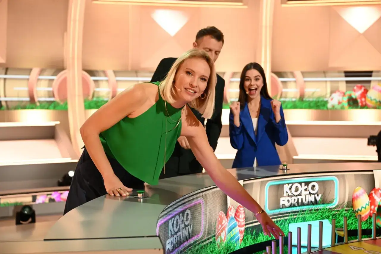 Trzy osoby na planie "Koła Fortuny" podczas emisji. Kobieta w zielonej bluzce naciska przycisk, druga w niebieskiej cieszy się z wygranej.