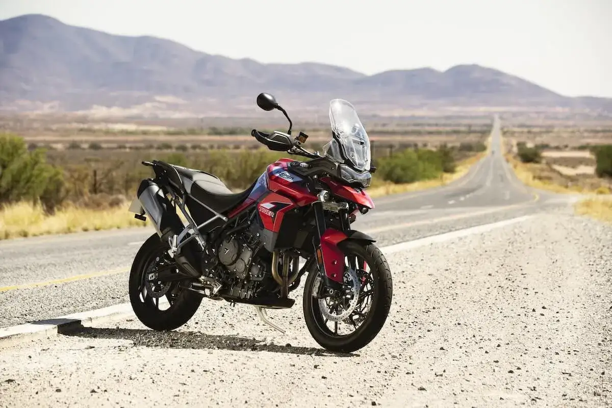 Czerwony motocykl Triumph Tiger 900 GT Pro zaparkowany na poboczu pustej drogi w krajobrazie z górami w tle.