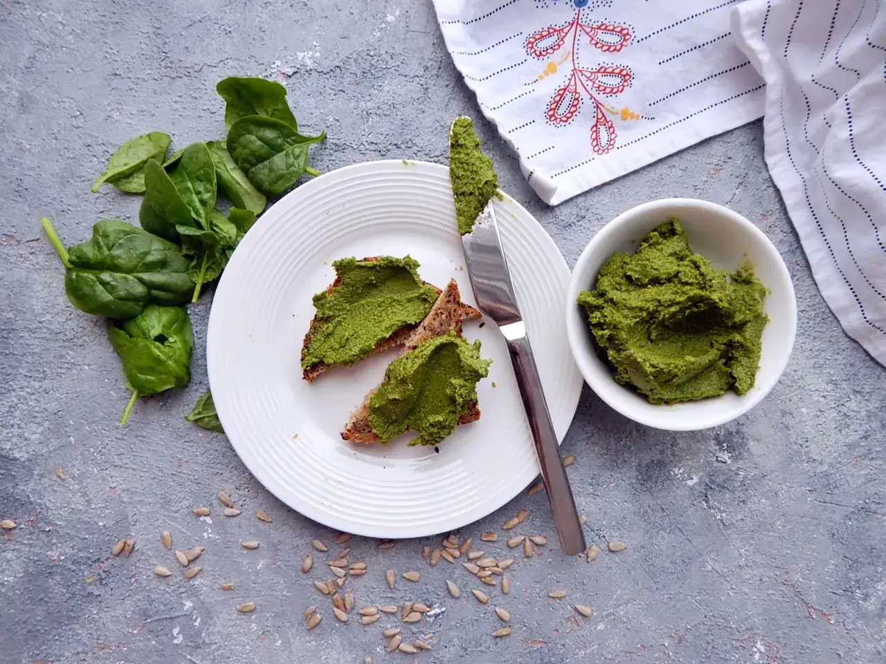 Pesto ze szpinaku do słoików na chlebie z pestkami słonecznika. Świeży szpinak obok talerza.