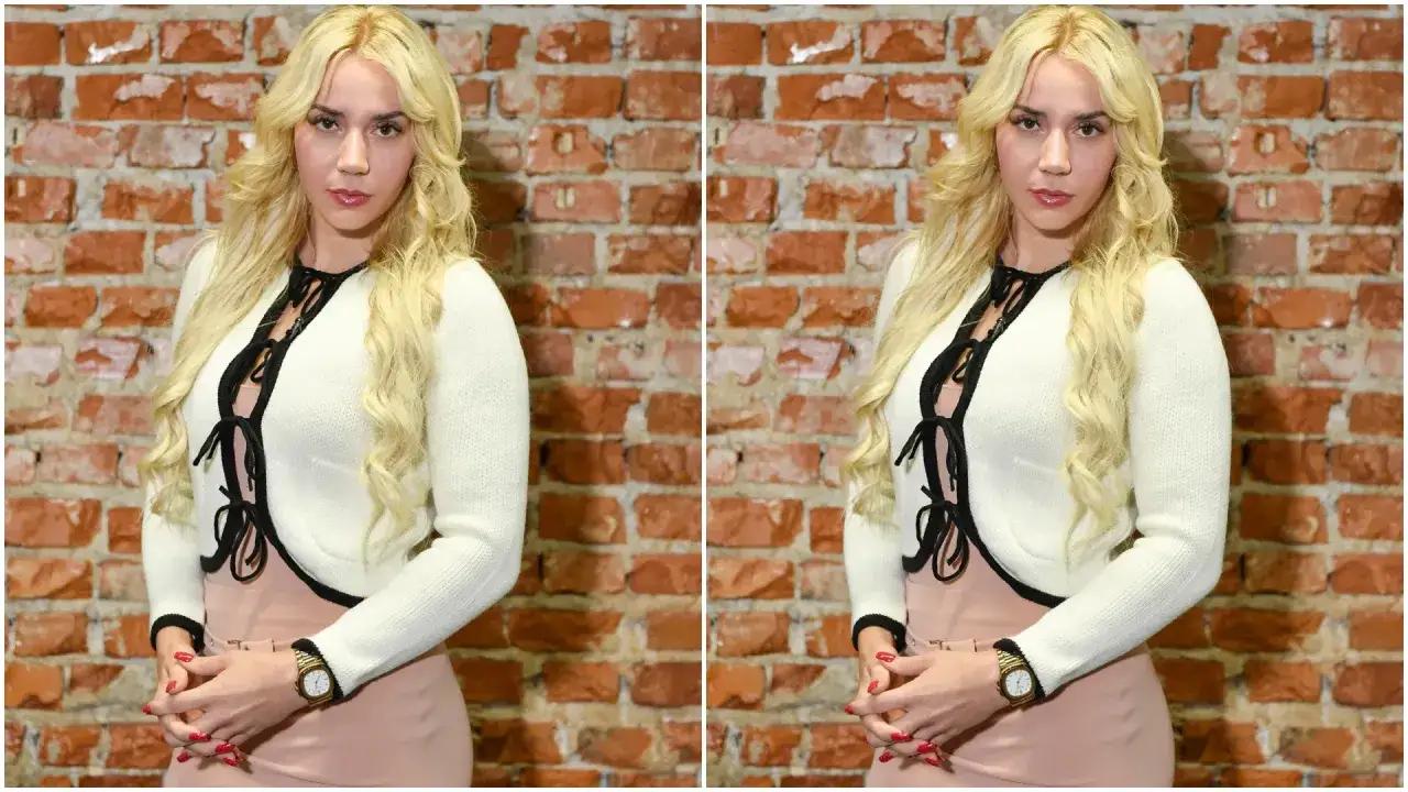 Marianna Schreiber na zdjęciach w białym sweterku i różowej sukience, z burzą blond włosów, pozuje na tle ceglanej ściany.