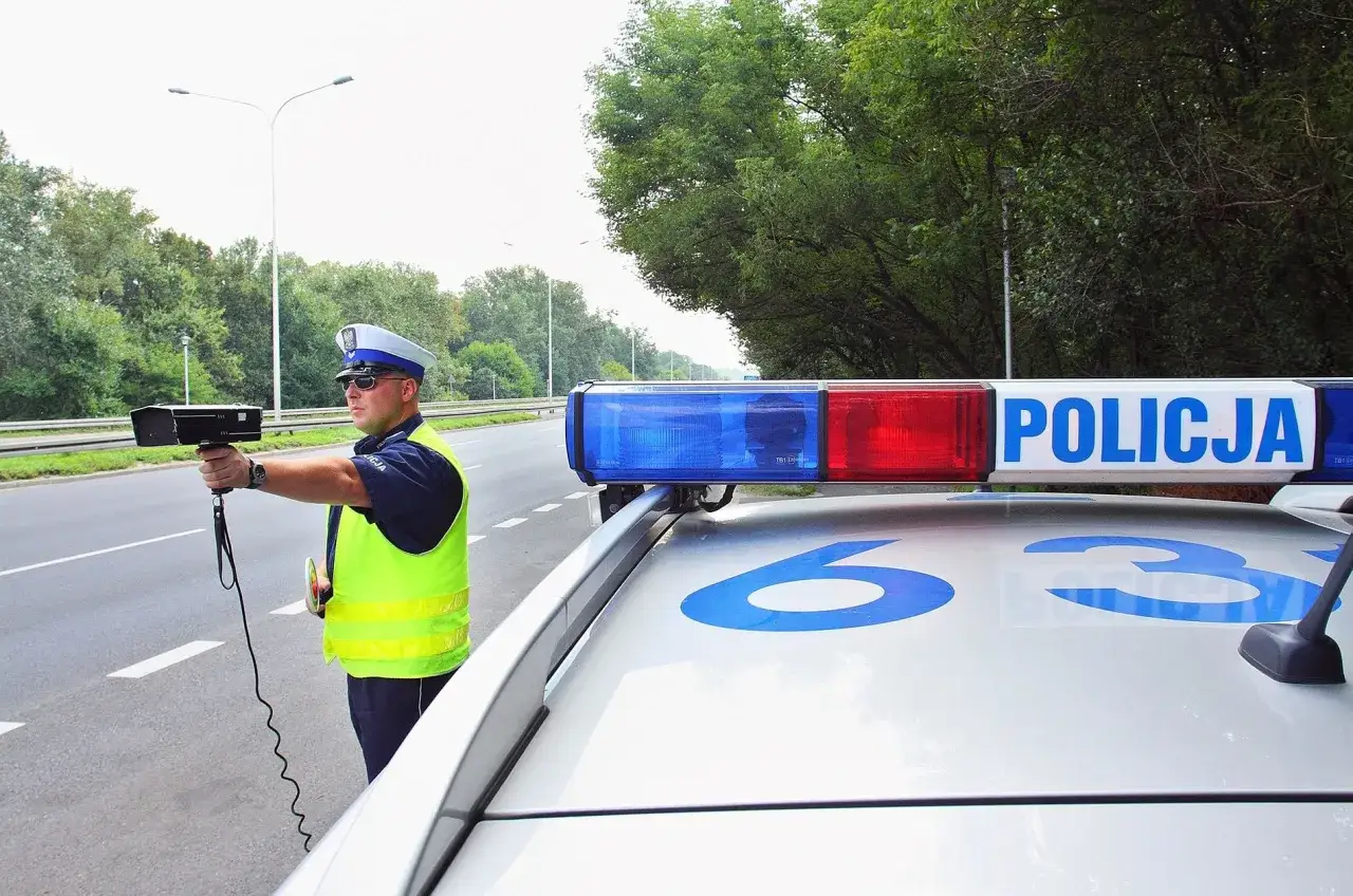 Policjant z radarem na drodze, obok radiowozu z napisem POLICJA.