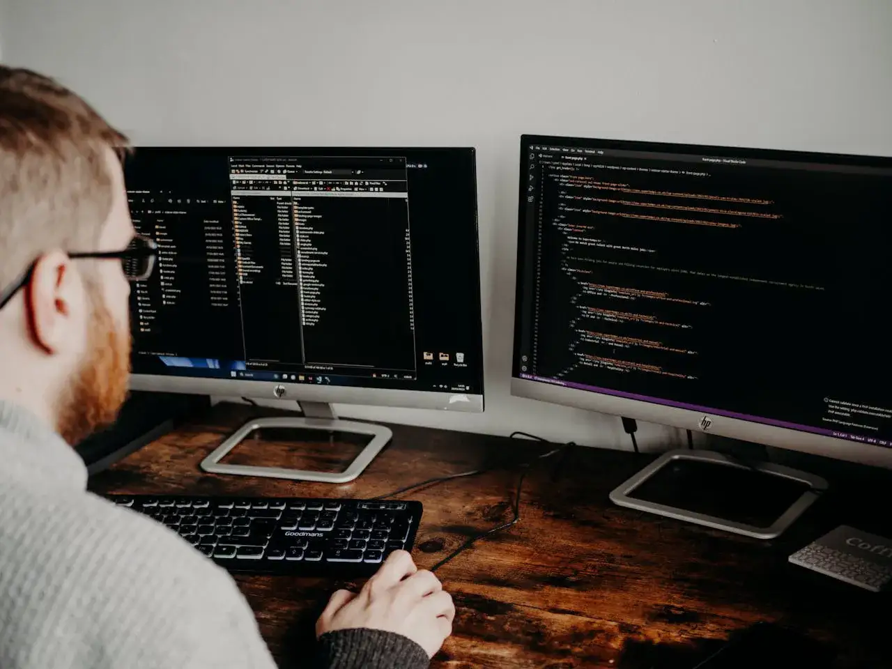 Mężczyzna z brodą i okularami pracuje przy dwóch monitorach z kodem. Czy szukanie pracy w IT bez doświadczenia, czy sam bootcamp wystarczy?