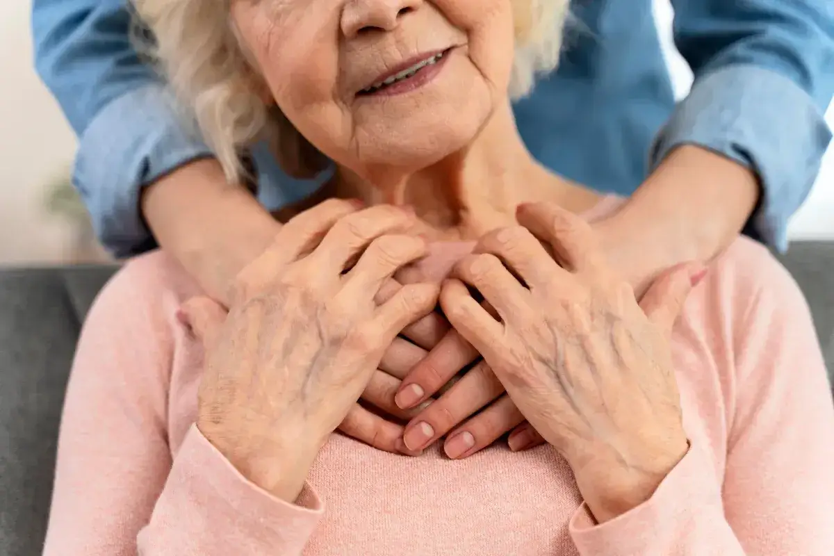 Dwie pary dłoni, starsza i młodsza, splatają się na piersi starszej kobiety. Obraz symbolizuje wsparcie i troskę, kluczowe przy ustalaniu, komu przysługuje zasiłek za opiekę nad osobą starszą.