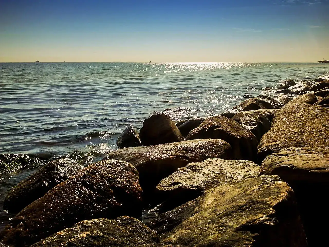 Kamienista plaża, fale rozbijające się o brzeg. W oddali widać statki i horyzont. Idealne miejsce na spacer w miejscowościach obok Ustki.