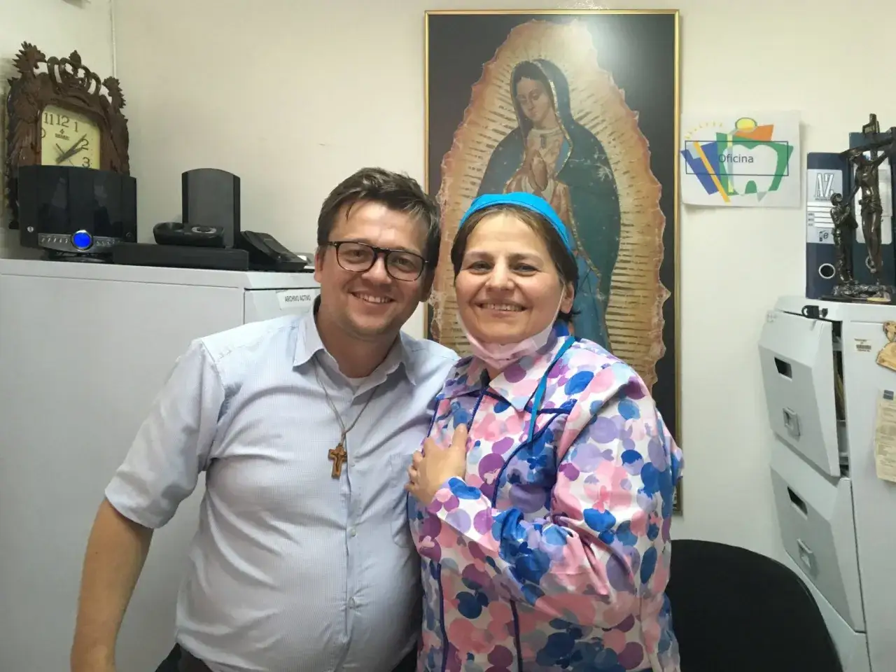 Mężczyzna w okularach i kobieta w kolorowym ubraniu pozują do zdjęcia na tle obrazu Matki Boskiej.