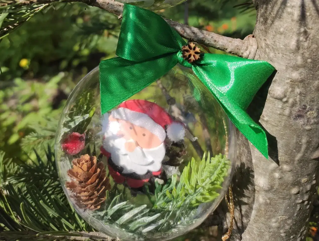 Przezroczysta bombka DIY z Mikołajem, szyszką i gałązkami, przewiązana zieloną wstążką, wisi na choince.