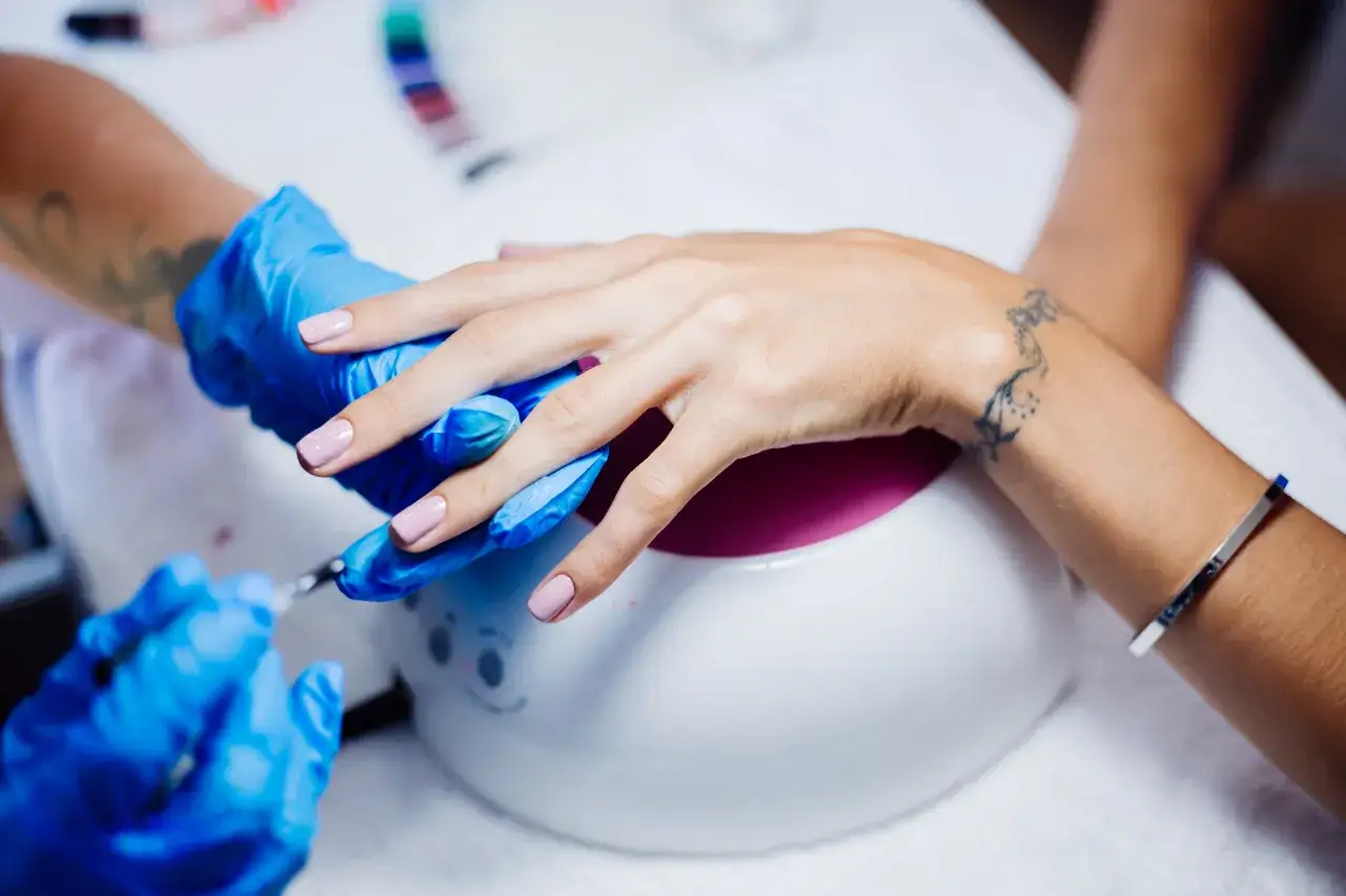 Manicure w trakcie: dłonie w niebieskich rękawiczkach, lampa UV, paznokcie żelowe. Kolejnosc robienia paznokci zelowych.