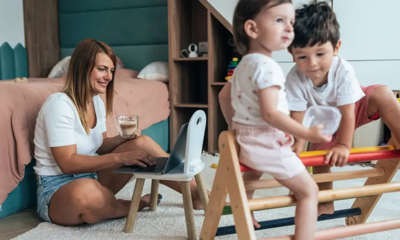 Kobieta pracuje na laptopie, pijąc kawę, podczas gdy dzieci bawią się obok. Pokazuje, jak pogodzić pracę z opieką nad dzieckiem.