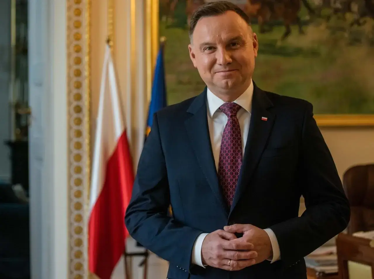 Prezydent Andrzej Duda w garniturze, z polską flagą w tle, czeka na moment, kiedy prezydent podpisze emerytury stażowe.