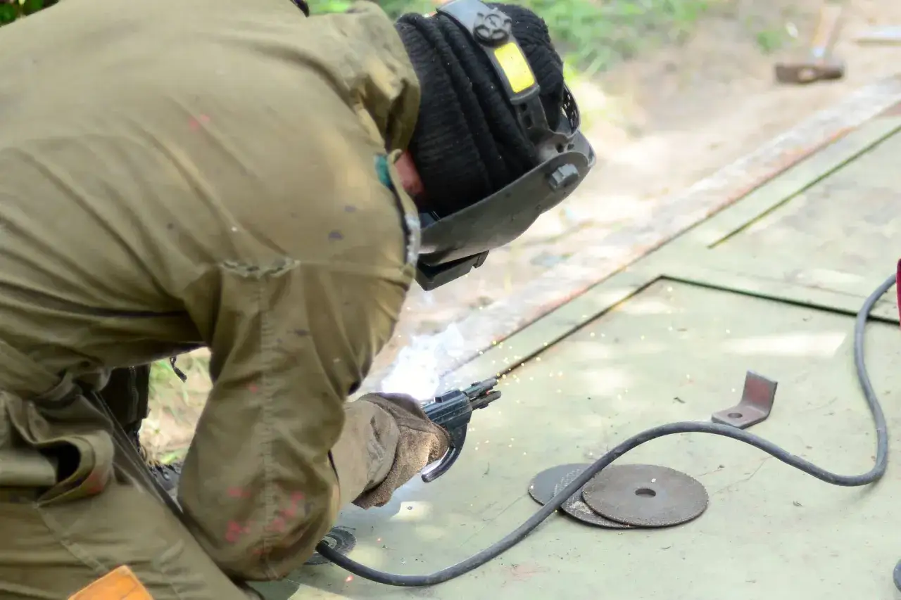 Spawacz w masce spawa metal, widać iskry i sprzęt.