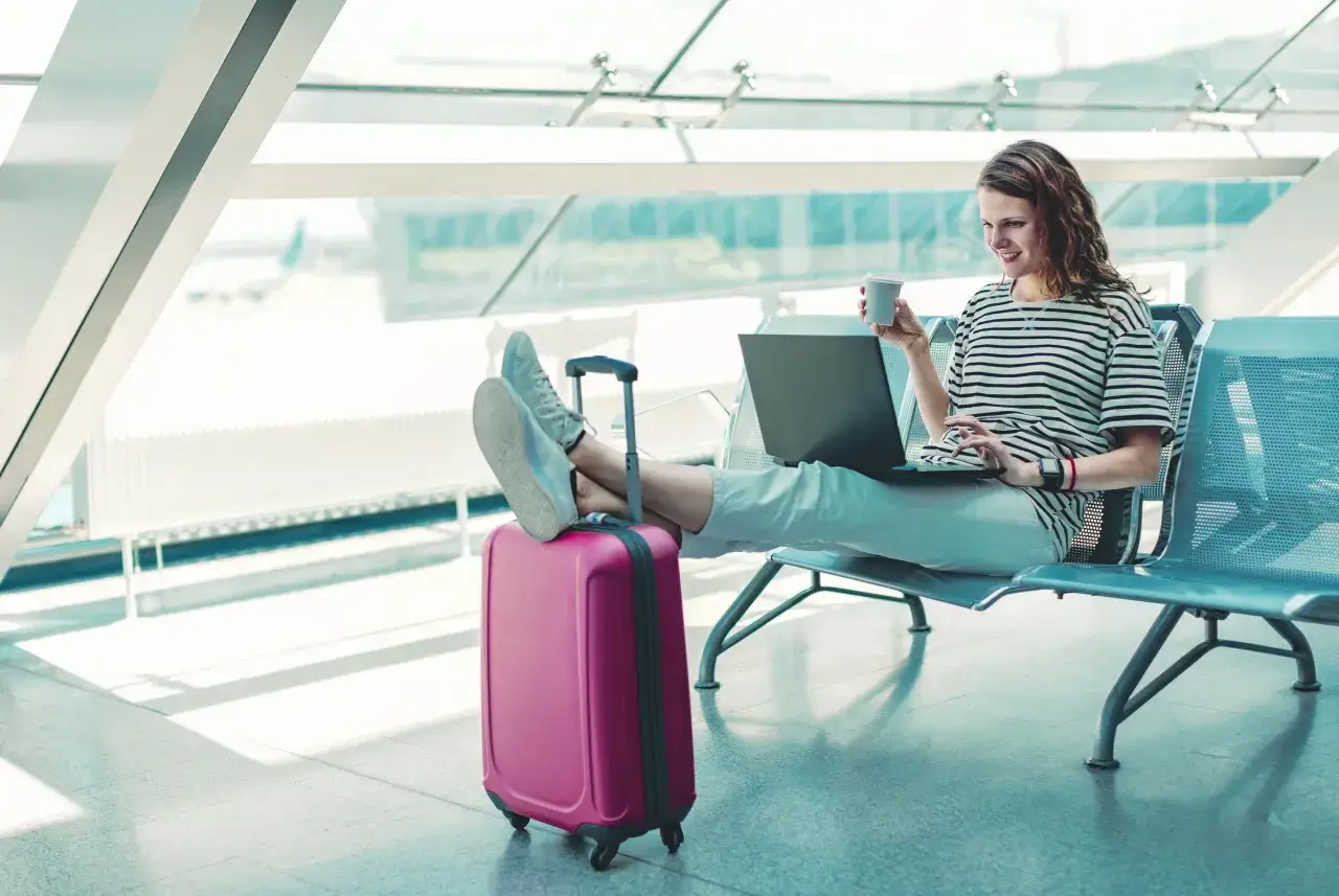 Kobieta z laptopem i kawą czeka na lotnisku, obok niej różowa walizka, idealna na to, co można zabrać do bagażu rejestrowanego.