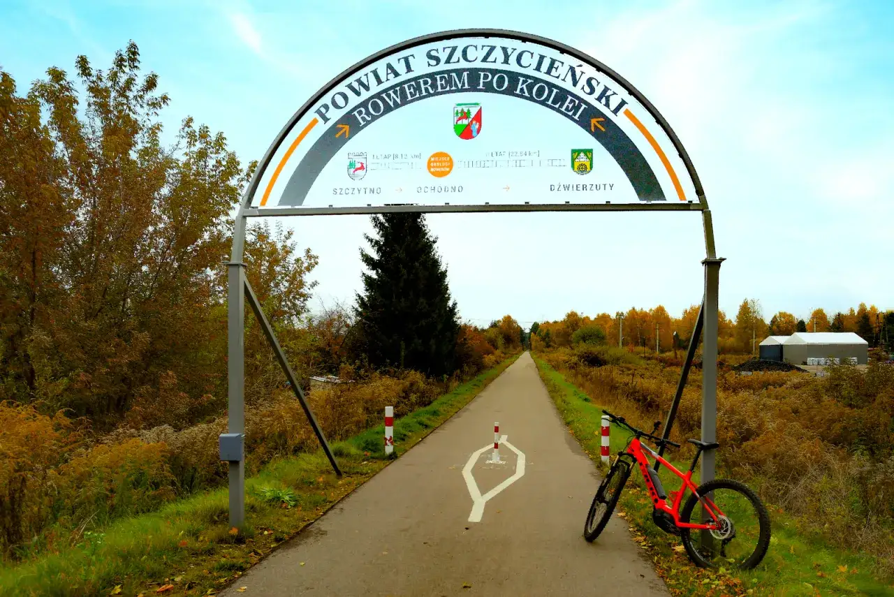 Pomarańczowy rower czeka na przygodę na trasie rowerowej w Szczytnie. Łuk nad drogą zaprasza do odkrywania powiatu.