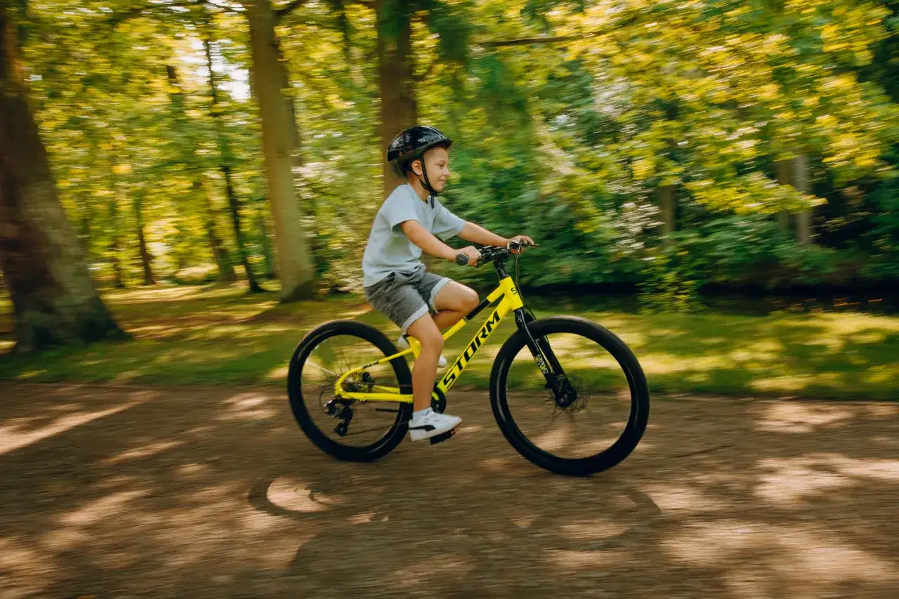 Chłopiec w kasku jedzie na żółtym rowerze po leśnej ścieżce.