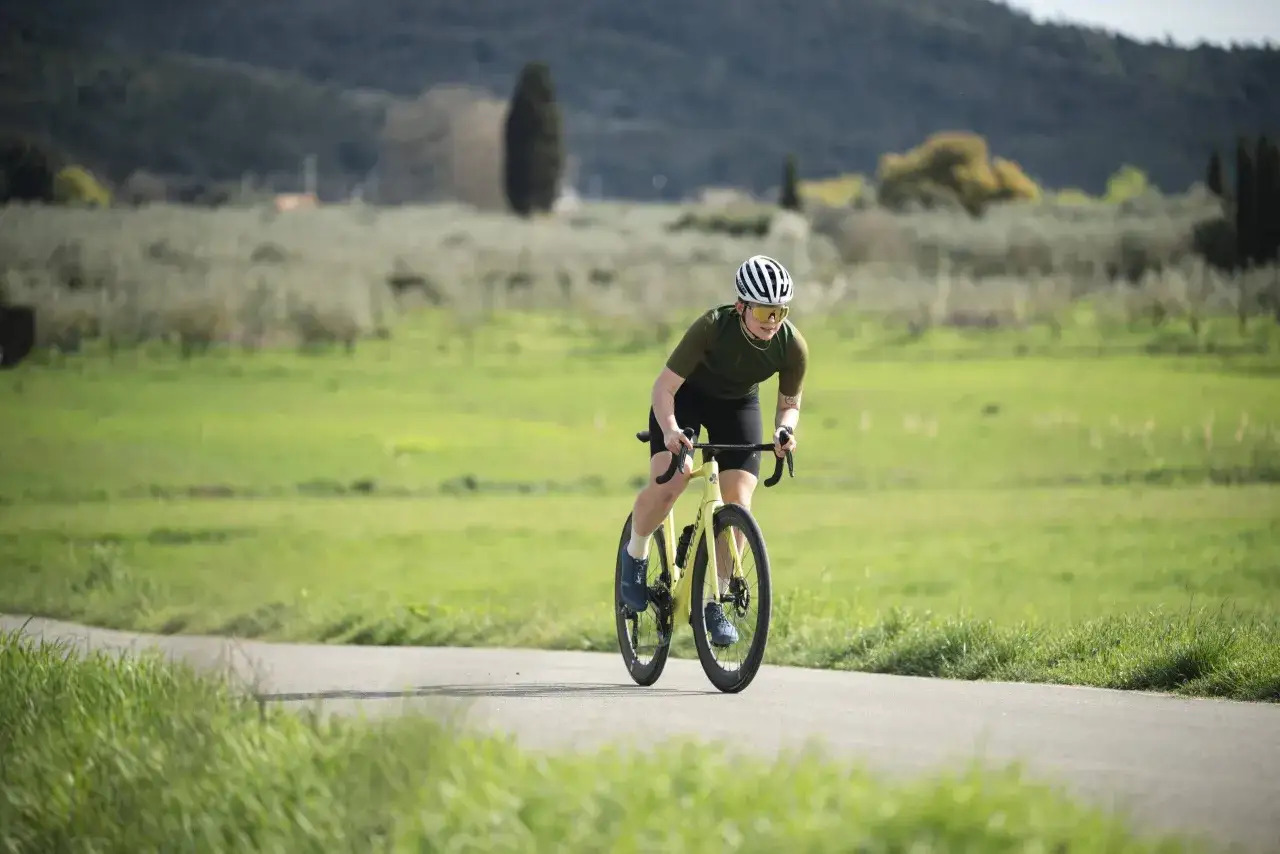 Kolarz w kasku i okularach ściga się po asfaltowej drodze. W tle zielone pola i wzgórza. To idealne miejsce na trening ftp kolarstwo.