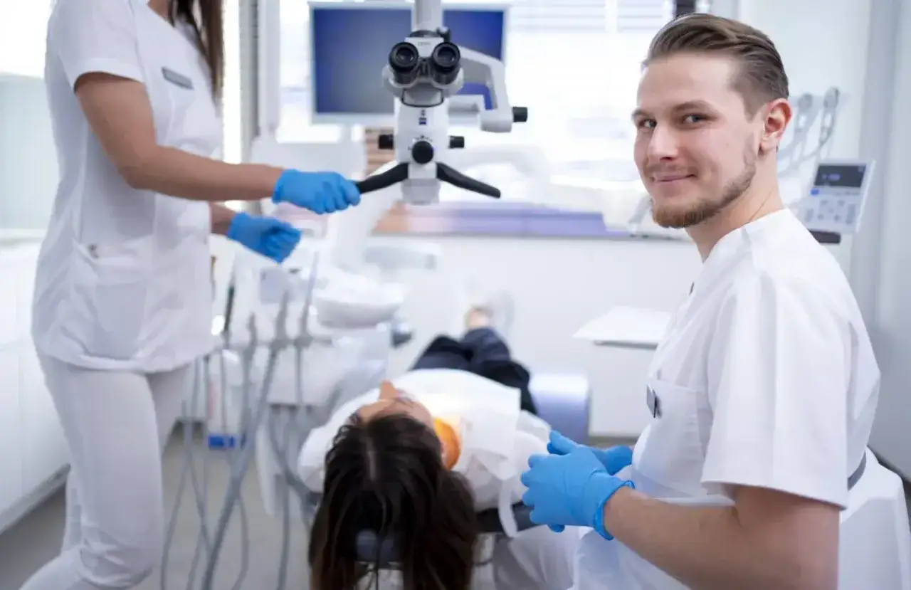 Uśmiechnięty dentysta w białym fartuchu i niebieskich rękawiczkach, pacjent na fotelu, asystentka przy mikroskopie.