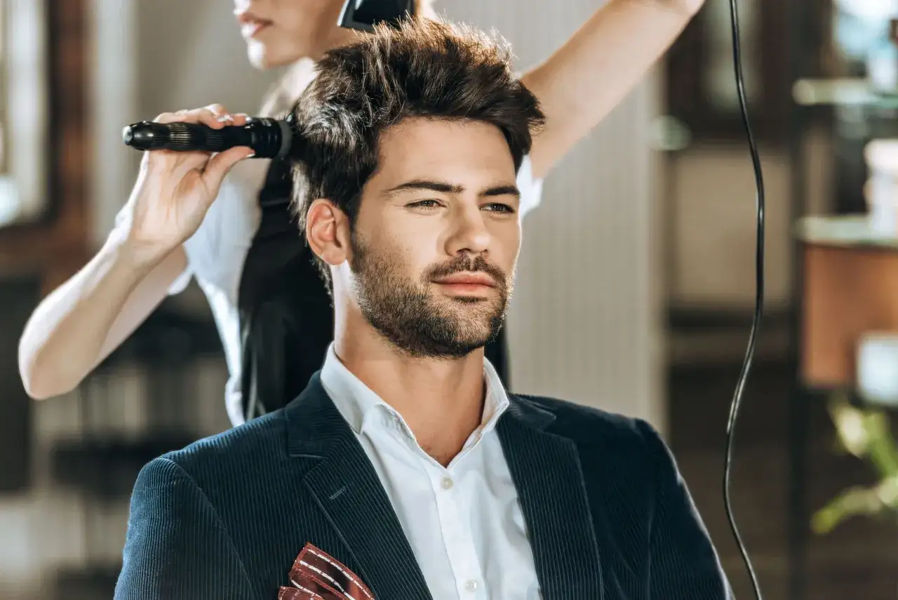 Mężczyzna w eleganckim garniturze czeka na stylizację fryzury męskiej. Fryzjerka suszy jego włosy suszarką.