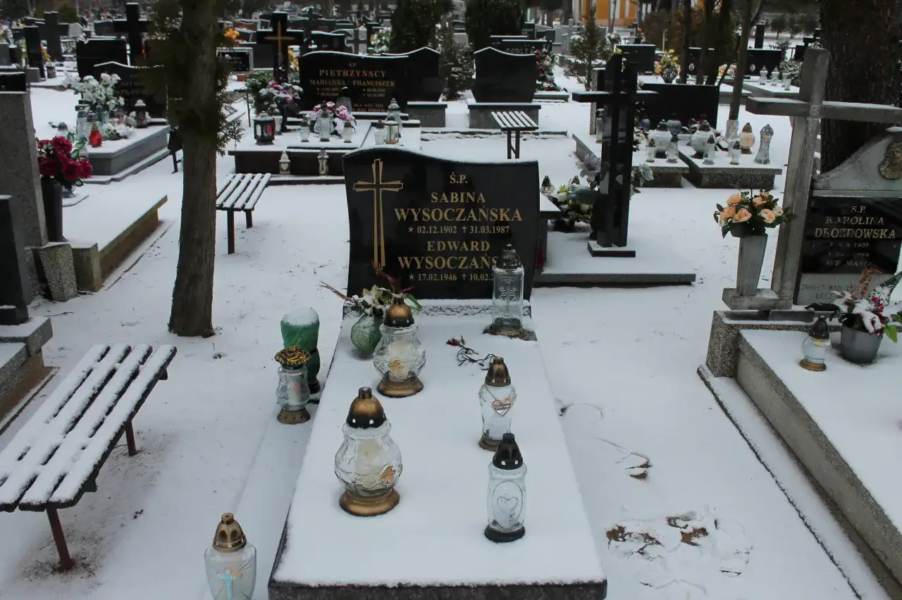 Zimowy cmentarz, grobonet krzyż wlkp. Śnieg przykrywa nagrobki i ławki. Płoną znicze, upamiętniając bliskich.