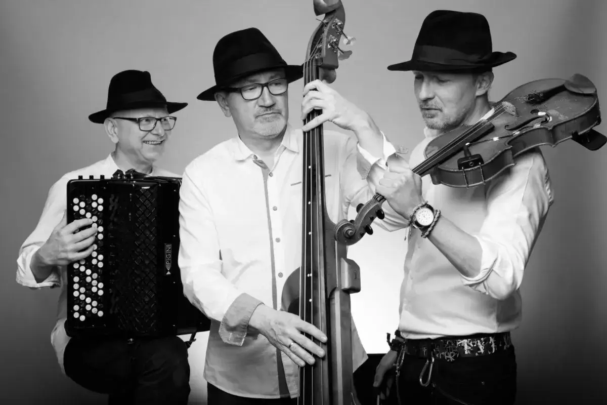 Trzech muzyków w czarnych kapeluszach grających muzykę klezmerską: akordeonista, basista i skrzypek.