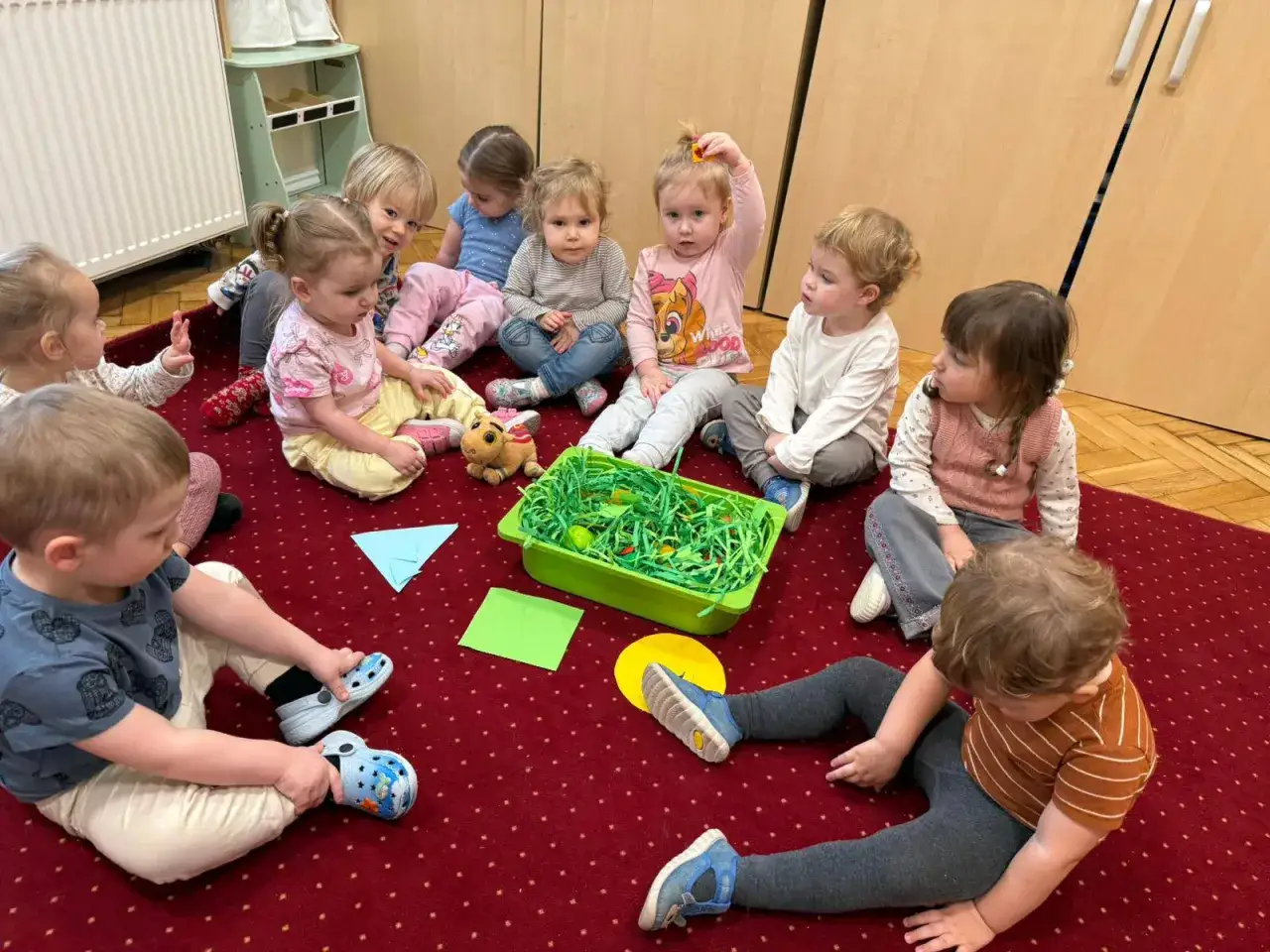 Dzieci siedzą na dywanie wokół zielonego pojemnika z zieloną trawą i jajkami.