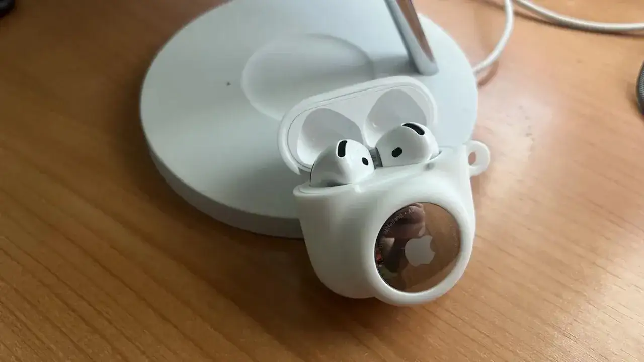 AirPods w białym etui z przyczepionym AirTagiem na drewnianym blacie.