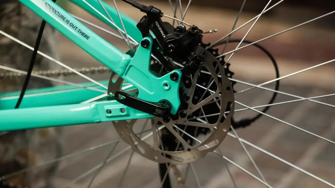 Tylna przerzutka i hamulec tarczowy na rowerze z turkusową ramą. Sztywna oś boost zapewnia stabilność podczas przygód.