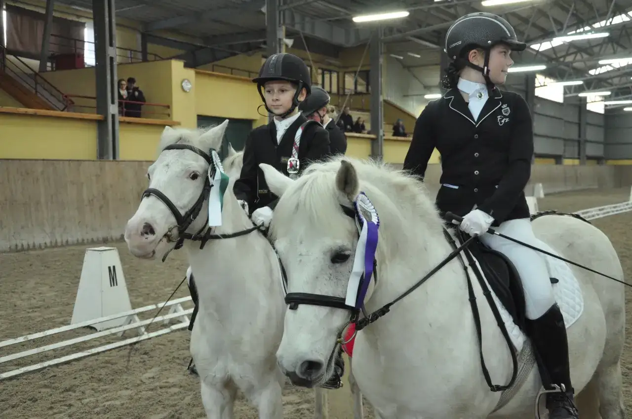 Dwie młode amazonki w strojach jeździeckich na białych koniach w hali ujeżdżalni.