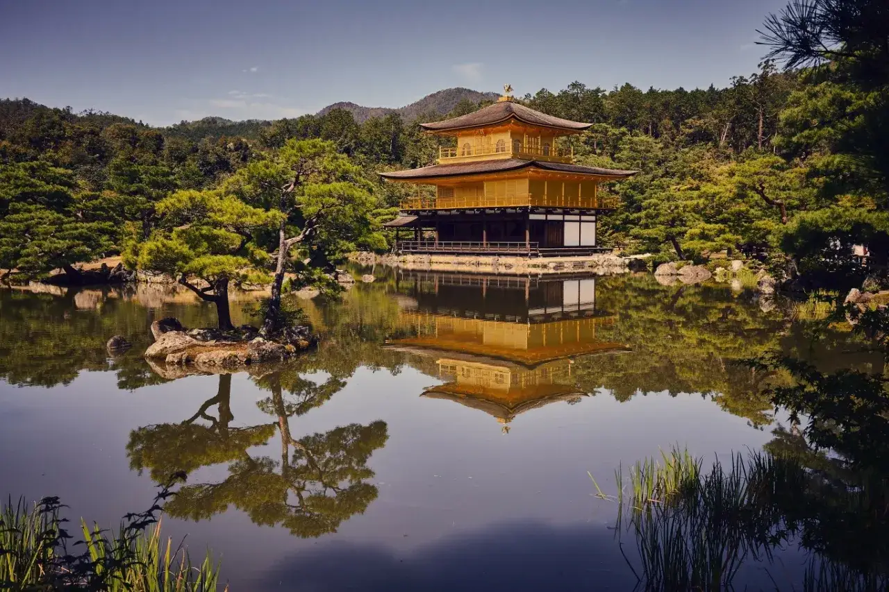 Złoty Pawilon Kinkaku-ji odbijający się w stawie, otoczony zielenią i drzewami.