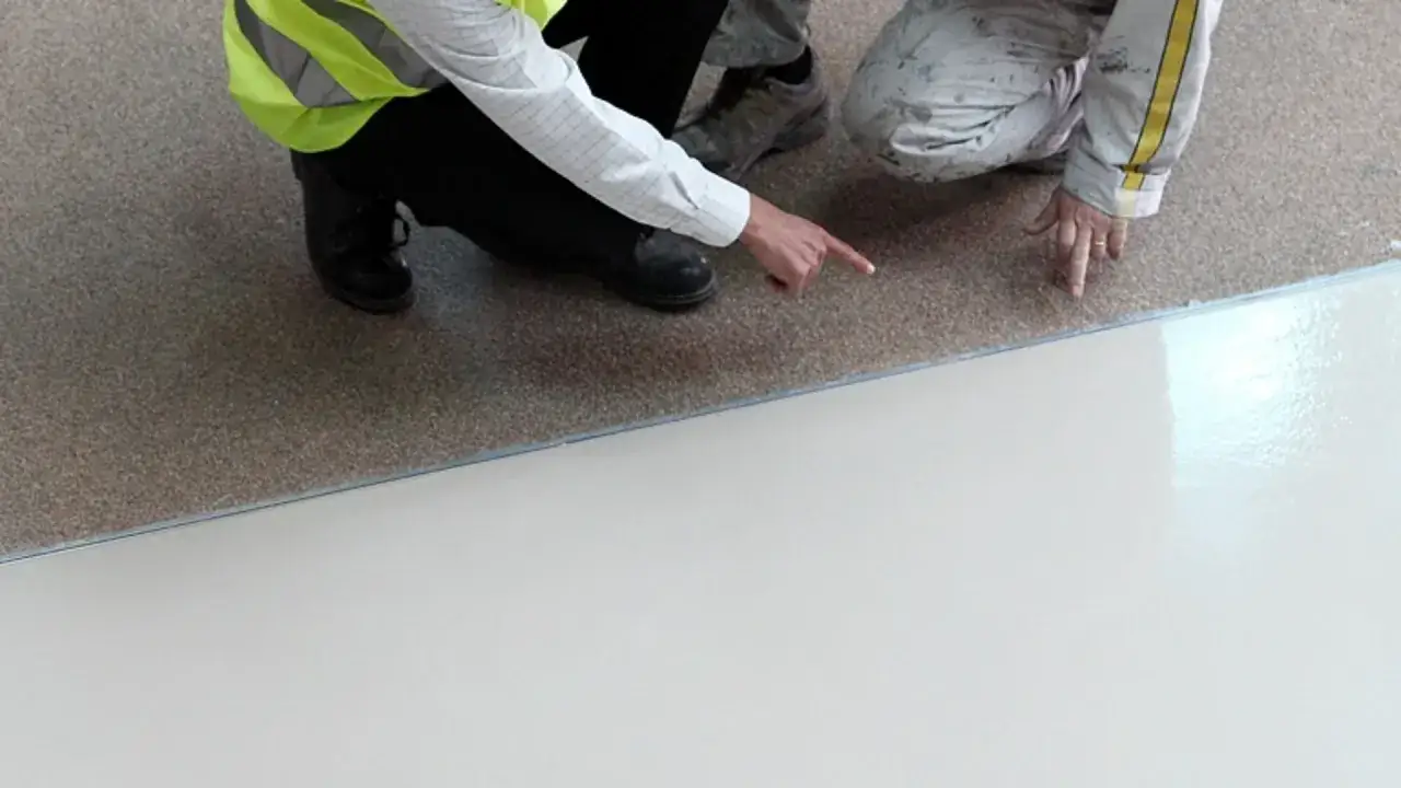 Dwóch pracowników sprawdza, do czego nie przywiera żywica epoksydowa. Jedna osoba w kamizelce odblaskowej wskazuje palcem na granicę między podłożem a nową posadzką.