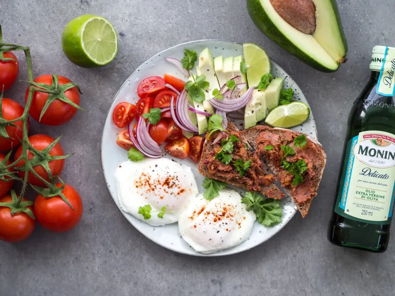 Talerz z jajkami sadzonymi, awokado, pomidorkami, cebulą i grzanką. Obok butelka oliwy.