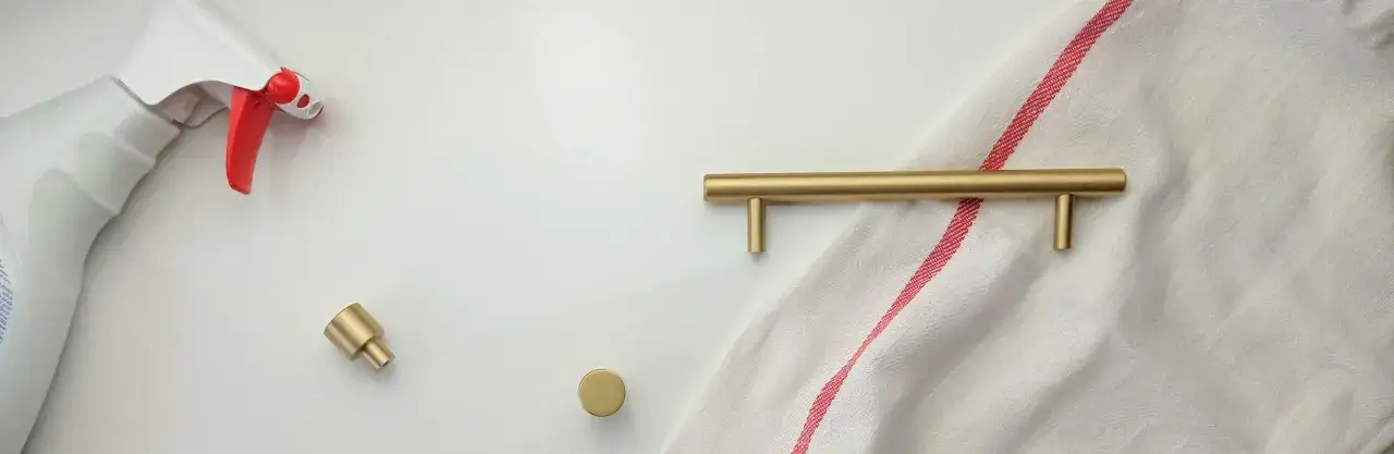 Biała butelka z płynem do czyszczenia, złote gałki i uchwyt meblowy na tle białej tkaniny w czerwone paski. Czy mosiądz rdzewieje? Nie, ale wymaga pielęgnacji.