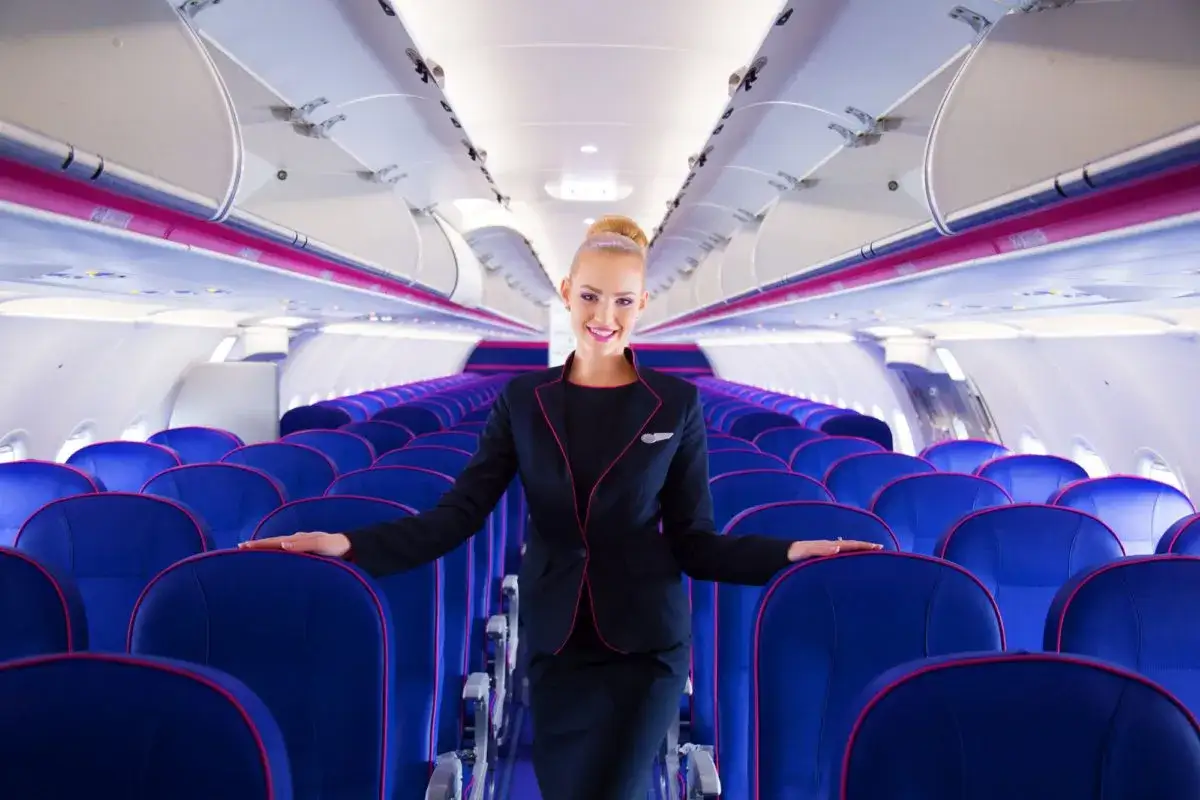 Uśmiechnięta stewardessa Wizz Air stoi w rzędach niebieskich foteli w samolocie.