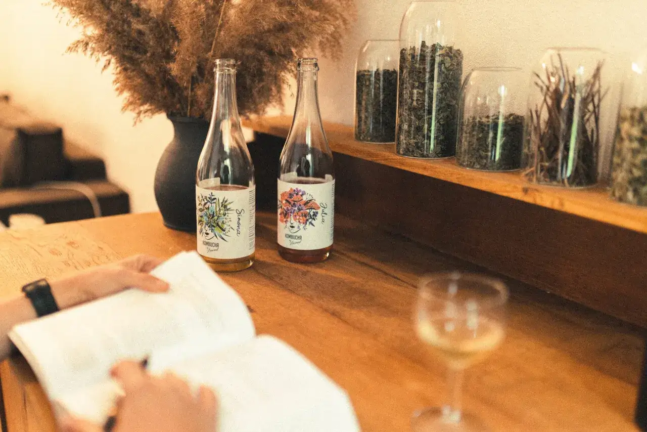 Dwie butelki kombuchy na drewnianym stole, obok kieliszek i otwarty notatnik.