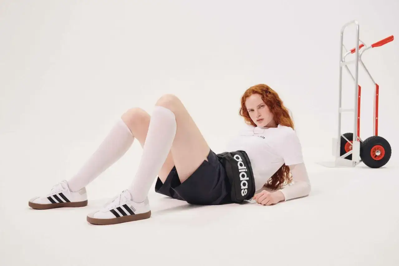 Rudowłosa modelka w białej koszulce i czarnych spodenkach z logo Adidas leży na podłodze. Czy Adidas ma zawyżoną rozmiarówkę?