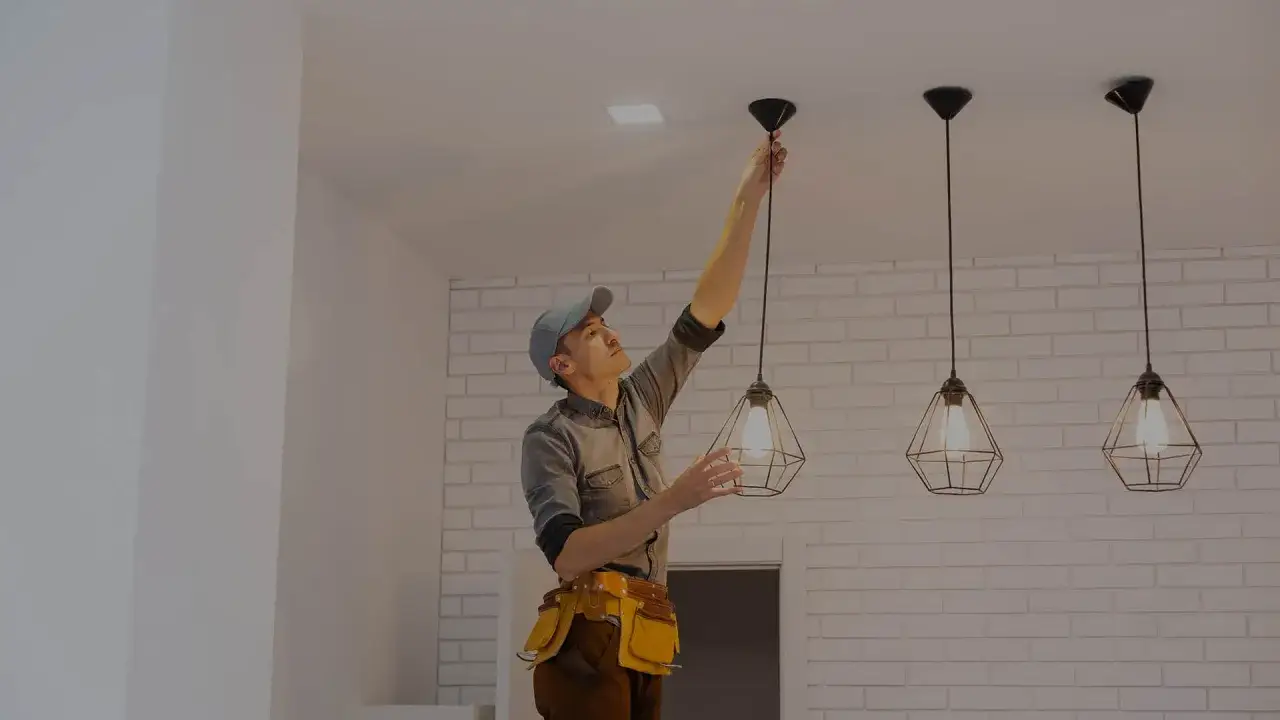 Elektryk w czapce i pasie z narzędziami montuje lampę wiszącą na suficie.
