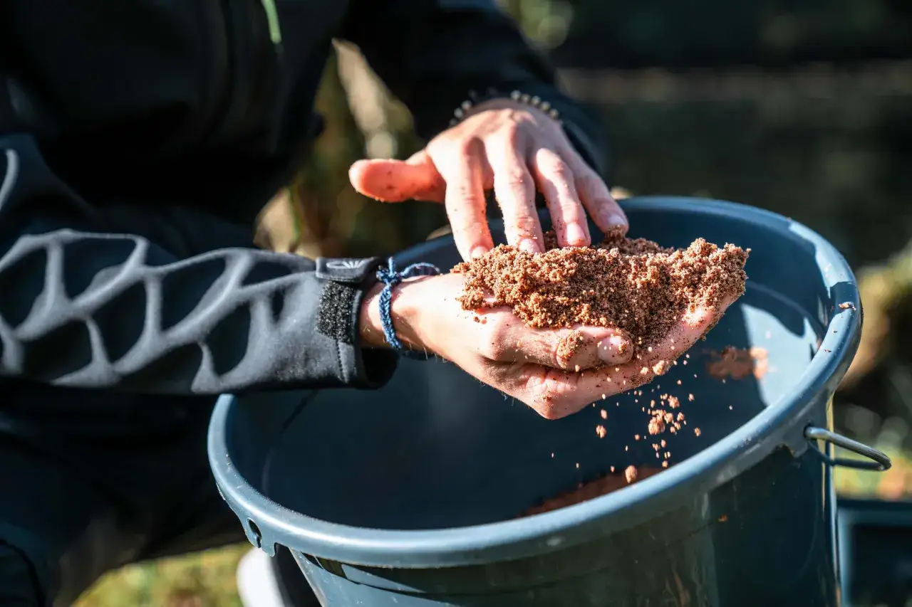 Ręce trzymają zanętę Traper Feeder nad wiaderkiem.