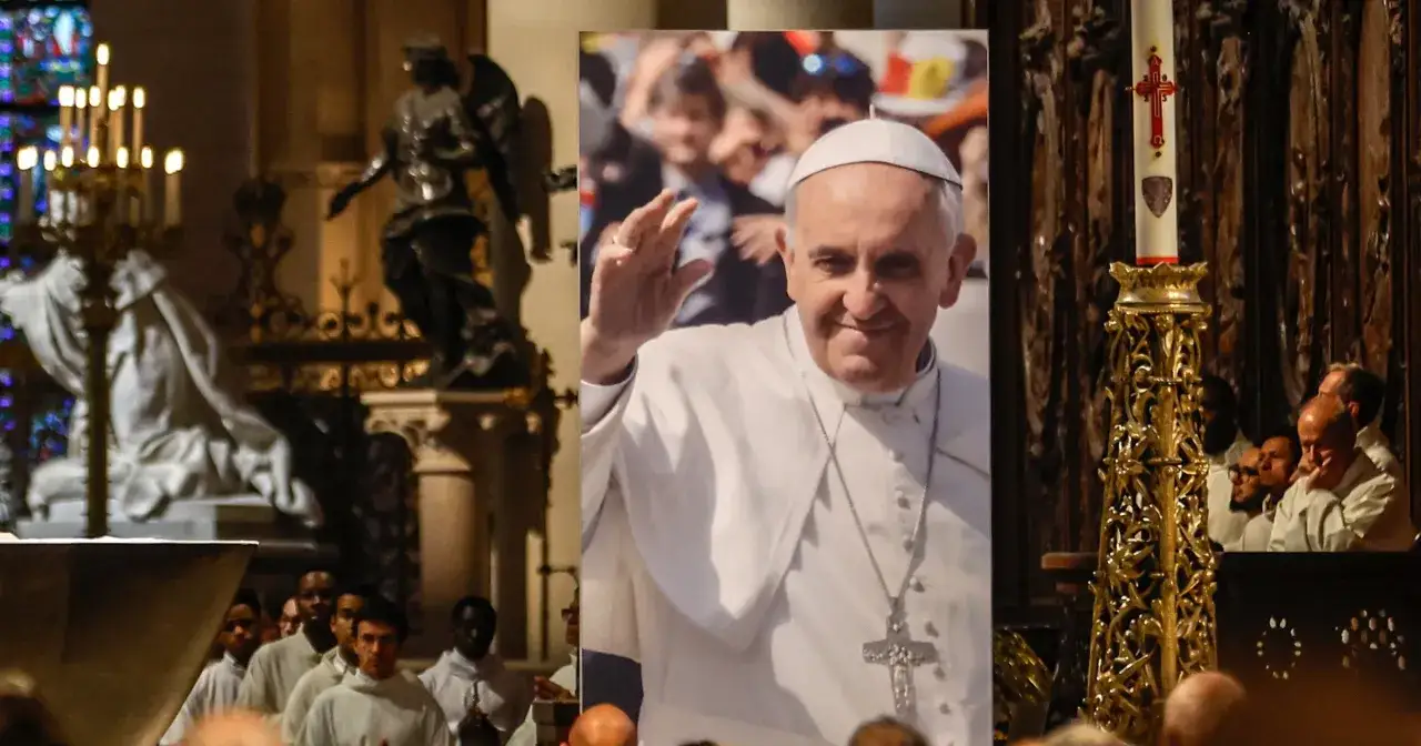 Papież Franciszek w białej szacie z krzyżem, na tle wnętrza kościoła.