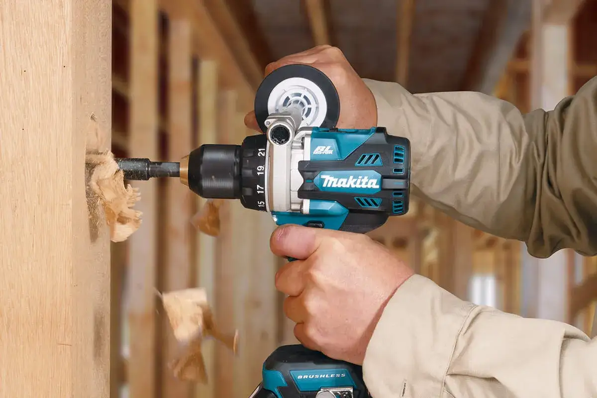 Dłoń trzyma wiertarkę Makita, która wierci otwór w drewnianej belce, wyrzucając wióry.